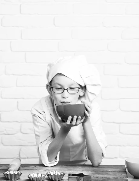 Jeune garçon petit chef cuisinier mignon en uniforme blanc et chapeau sur le visage drôle avec des lunettes debout près de la table avec des moules à rouleau pour gâteaux tenant bol rouge et souffler dans la farine sur fond de mur de brique — Photo