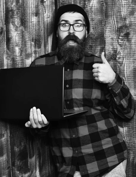 Γενειοφόρος άνδρας, μακριά γενειάδα. Βάναυση Καυκάσιος έκπληκτος αξύριστος hipster κρατώντας το laptop στο κόκκινο μαύρο καρώ πουκάμισο με καπέλο και γυαλιά σε backgroun καφέ vintage ξύλινα στούντιο — Φωτογραφία Αρχείου