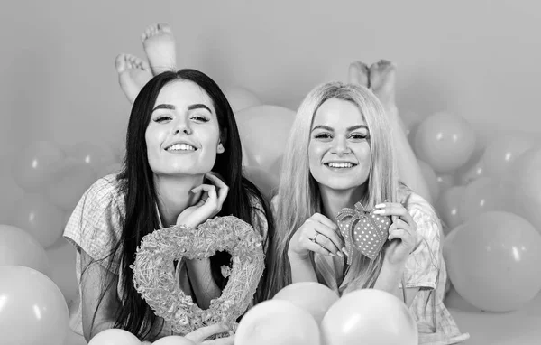 Meisjes lag in de buurt van ballonnen, houdt hart speelgoed, roze achtergrond. Zusters, vrienden in pyjama in pyjama's partij. Blonde en brunette op de lachende gezichten droomt over liefde en datum. Valentines day concept — Stockfoto