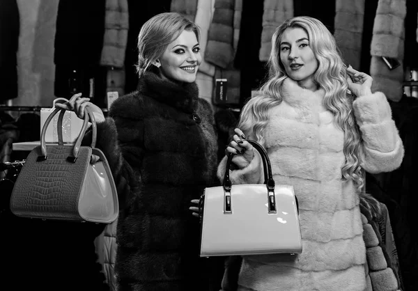 Vrouwen in bontjassen met zakken in bont winkel. Winter kleding en glamour concept. Dames met make-up winkelen in mode. Meisjes met lachende gezichten in zwart-wit bontjassen houden portemonnees in handen — Stockfoto