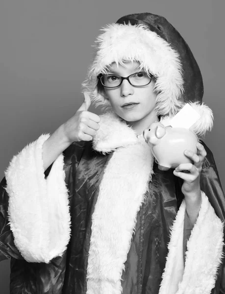 Młody chłopak ładny Święty Mikołaj z okulary w czerwonym swetrze i kapelusz Boże Narodzenie nowy rok różowy świnia piggy bank gospodarstwa i pokazywanie chłodnym na tle niebieski studio — Zdjęcie stockowe