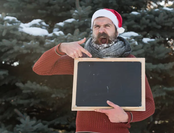 Schreeuwen man in Kerstman hoed, hipster met baard en snor bedekt met white frost, punten tijdens lege zwarte board of schoolbord op winterdag op natuurlijke achtergrond, kopiëren vriendelij — Stockfoto