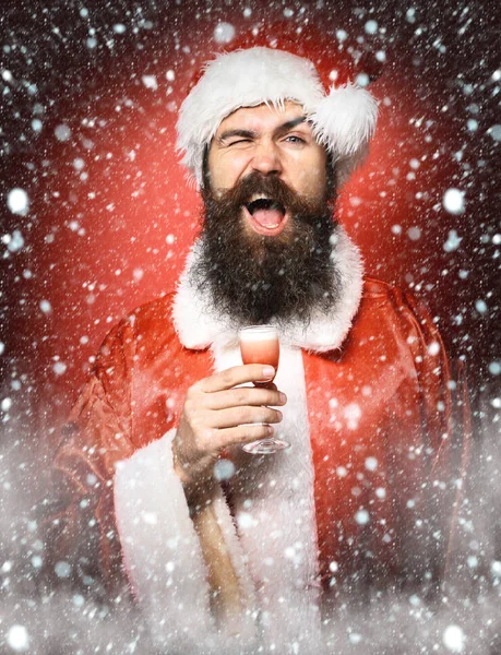 Guapo barbudo hombre de Santa Claus con barba larga en la cara divertida sosteniendo vaso de trago alcohólico en navidad o suéter de Navidad y sombrero de año nuevo en fondo rojo estudio —  Fotos de Stock