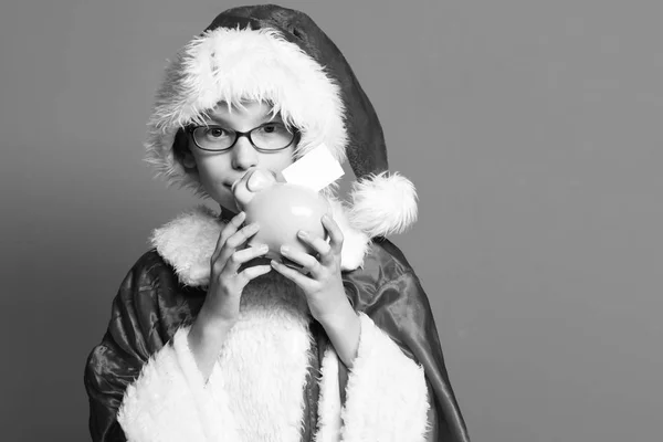年轻可爱的圣诞老人男孩与眼镜在圣诞毛衣和新年圣诞帽举行和亲吻粉红色的猪猪银行在红色的工作室背景, 复制垃圾 — 图库照片