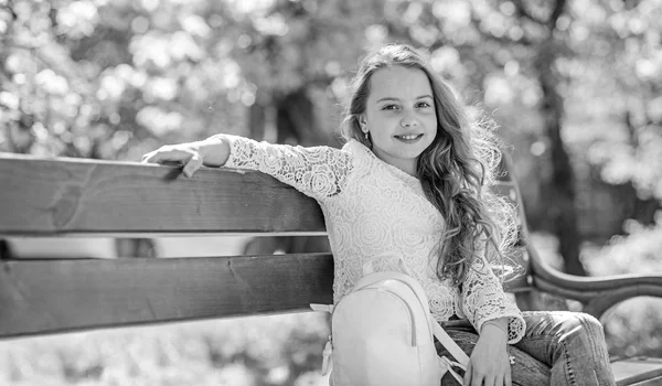 Fille sur le visage souriant s'assoit sur le banc, sakura sur le fond, déconcentré. Fille se détendre tout en marchant dans le parc près de la fleur de cerisier. Concept d'accessoires mode. Enfant mignon avec sac à dos profiter ensoleillé jour de printemps — Photo