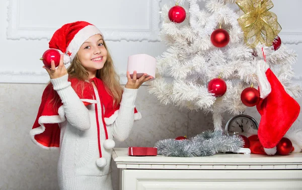 Child decorating christmas tree with red balls ornaments. Cherished holiday activity. Kid in santa hat decorating christmas tree. Family tradition concept. Girl kid decorating christmas tree