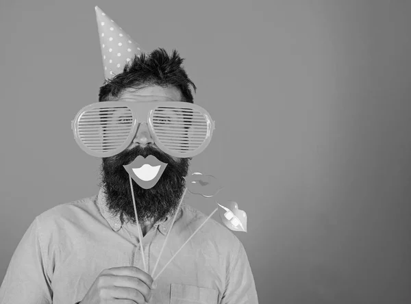 O tipo de chapéu de festa festeja, posando com adereços fotográficos. Hipster em óculos de sol gigantes comemorando. Homem com barba na cara alegre mantém lábios sorridentes em paus, fundo vermelho. Conceito de diversidade emocional — Fotografia de Stock