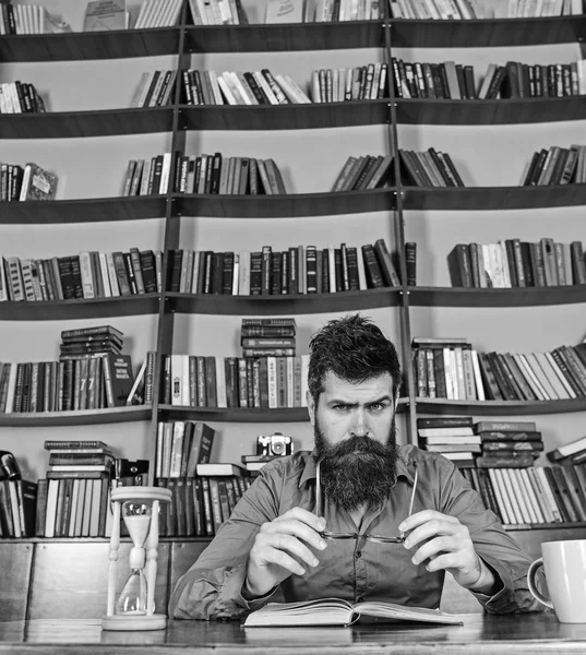 Man on busy face reading book, bookshelves on background. Education and science concept. Scientist sits at table with eyeglasses and hourglass. Teacher or student with beard studying in library — Stock Photo, Image