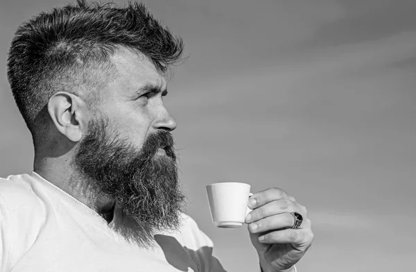 Man with long beard enjoy coffee. Coffee gourmet concept. Man with beard and mustache on strict face drinks coffee, blue sky background, defocused. Bearded man with espresso mug, drinks coffee