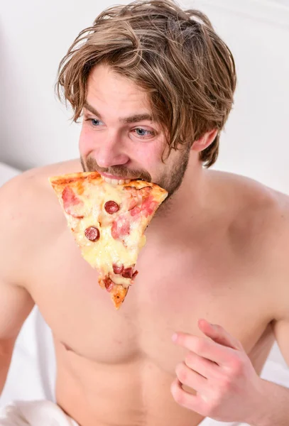 Man bearded handsome bachelor eating cheesy food for breakfast in bed. Handsome man holds a piece of pizza in his hands and is about to eat it. Weekend. — Stock Photo, Image