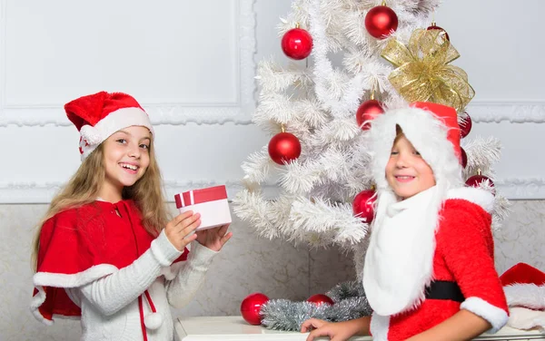 Rozpakowanie prezenty świąteczne. Wesołych Świąt Bożego Narodzenia. Chłopiec dziecko ubrane jak santa z białą brodą sztuczne i red hat dać pudełko do dziewczyny. Dzieci świętować Boże Narodzenie prezenty. Santa przynieść prezenty — Zdjęcie stockowe