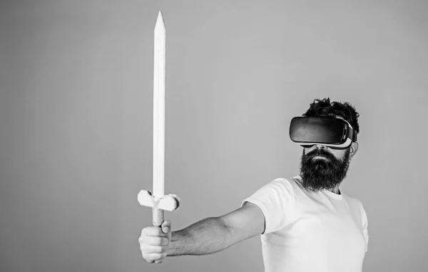 Gamer concept. Guy with head mounted display holds sword, play fighting game in VR. Man with beard in VR glasses, light blue background. Hipster on serious face enjoy play game in virtual reality
