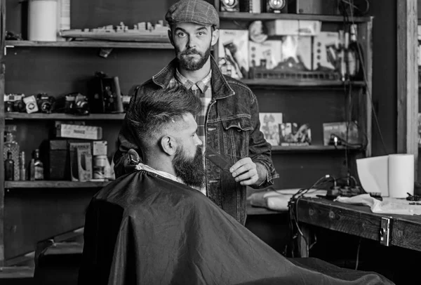 Concepto de aseo. Peluquero ocupado con la barba de aseo de cliente hipster, suministros de belleza en el fondo. Hipster con barba cubierta con capa que sirve por peluquero profesional en la elegante barbería — Foto de Stock