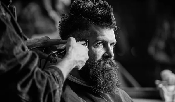 Hands of barber with hair clipper, close up. Hipster bearded client getting hairstyle. Barbershop concept. Barber works with hair clipper. Man with beard in hairdressers chair, dark background.