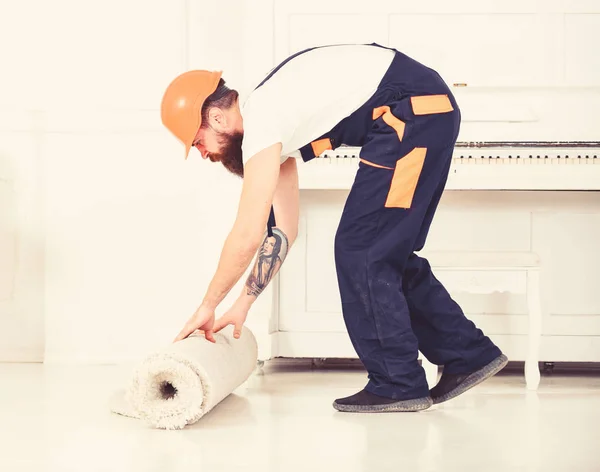 Relocating concept. Man with beard, worker in overalls and helmet rolling carpet, white background. Loader wrappes carpet into roll. Courier delivers furniture in case of move out, relocation