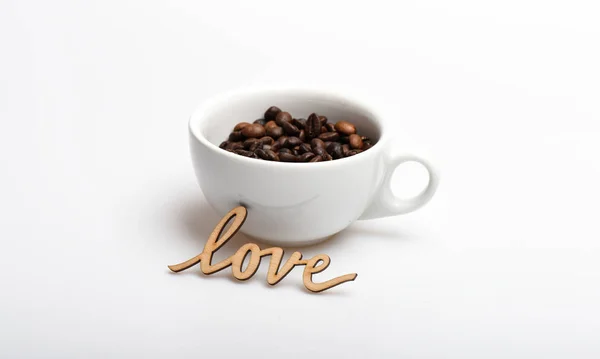 Романтична кава-брейк. Кавові зерна на білому тлі. Насолоджуйтесь кавовим напоєм. Дата в концепції кафе. Напій складається з кофеїну. Керамічна чашка з кавовими зернами та дерев'яним словом любов — стокове фото