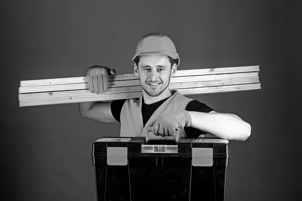 Muž v helmu, přilbu drží nástrojů a dřevěné trámy, šedé pozadí. Tesař, dělník, tvůrce, truhlář na šťastný obličej nese trámy na rameni. Profesionální truhlář koncept — Stock fotografie
