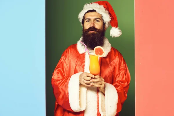 Handsome bearded santa claus man with long beard on serious face holding glass of nonalcoholic cocktail in christmas or xmas sweater and new year hat on colorful studio background — Stock Photo, Image