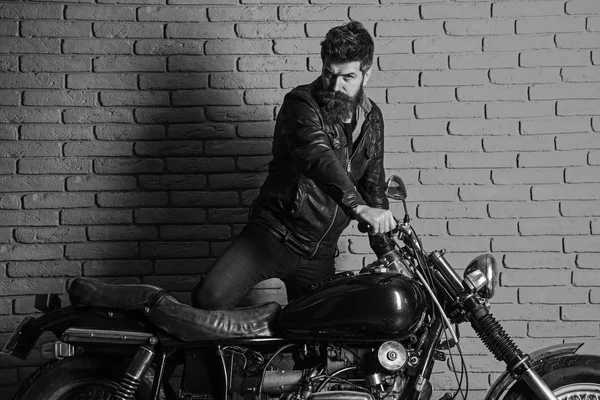 Мужская страсть. Мужчина с бородой, байкер в кожаной куртке возле мотоцикла в гараже, кирпичный настенный фон. Хипстер, жестокий байкер на серьезном лице в кожаной куртке садится на мотоцикл — стоковое фото