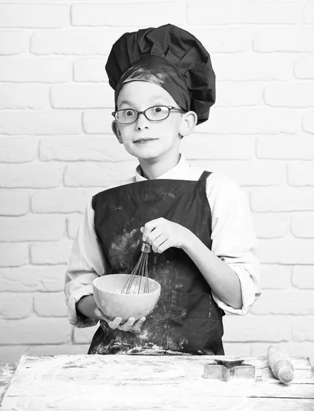 Young boy cute cook chef in red uniform and hat on stained face flour with glasses standing near table with rolling pin and cooking with kitchen whisk in turquoise bowl on white brick wall background — Stock Photo, Image