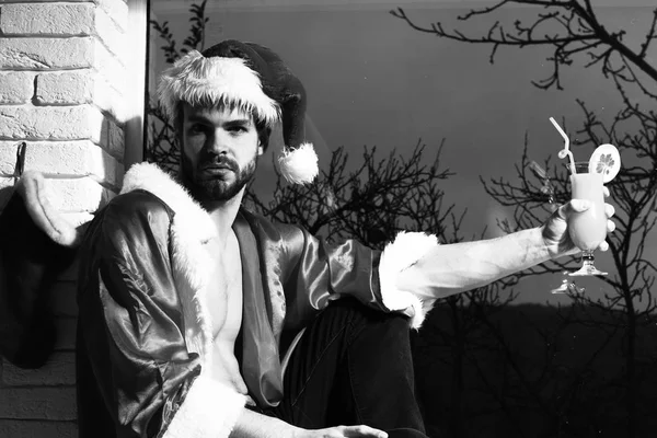 Genç yakışıklı sakallı seksi Noel Baba şapkalı ve ceketli kaslı çıplak gövdeli adam pencere kenarında oturuyor ve siyah ve tuğla duvar arkasında alkollü kokteyl bardağı tutuyor. — Stok fotoğraf