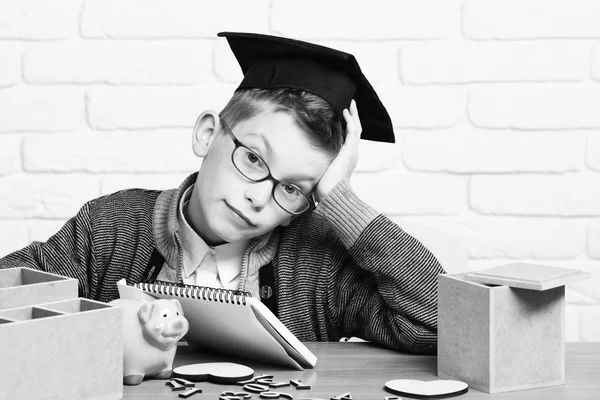 Ung söt elev pojke i grå tröja och glasögon sitter vid skrivbordet med copybook trä siffror rosa gris bank i examen mössa i klassrummet på vit tegelvägg bakgrund — Stockfoto