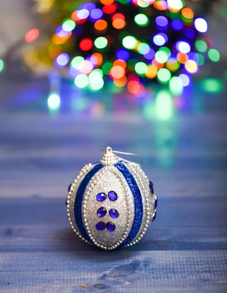 Christmas ornaments concept. Ball ornament on blue wooden surface. Pick colorful decor for christmas tree. Christmas ornament single ball on defocused garland colorful lights background
