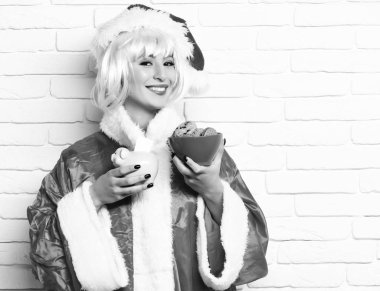 oldukça sevimli seksi Noel Baba kız ya da pembe peruk içinde yeni yıl kırmızı kazak ve Noel şapkalı gülümseyen esmer kadın domuz domuz kase beyaz tuğla duvar zemin üzerine banka ve çikolata kalça tanımlama bilgilerini tutar