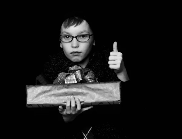 Small boy or cute nerd kid in glasses, hat and fashionable knitted scarf on black background holds red present box with golden bow and thumb u — Stock Photo, Image