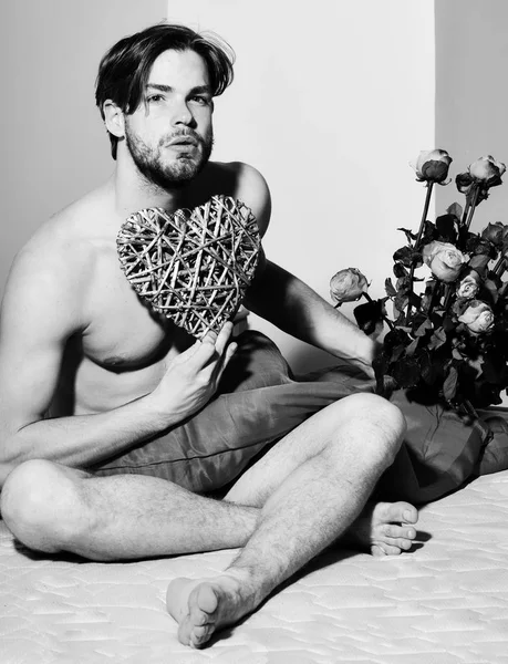 Joven guapo barbudo sexy macho hombre con elegante barba sentado desnudo en la cama bajo la manta roja celebración ramo de rosas naranjas y decorativo San Valentín escuchar — Foto de Stock