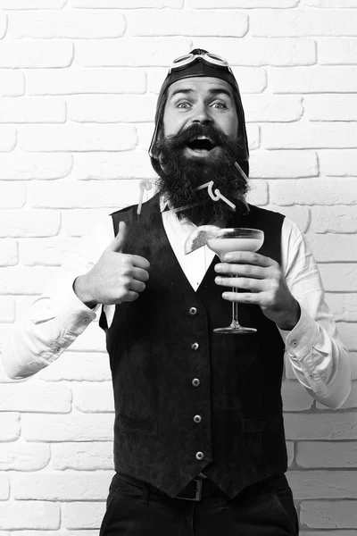 Hombre aviador barbudo guapo con barba larga y bigote en la cara feliz sosteniendo vaso de cóctel alcohólico en chaleco de cuero de gamuza vintage con sombrero y gafas sobre fondo de pared de ladrillo blanco —  Fotos de Stock