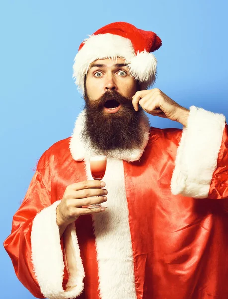 Όμορφος γενειοφόρος άνδρας Βασίλη με μακριά γενειάδα στο έκπληκτος πρόσωπο κρατά ποτήρι αλκοολικού βολή στο Χριστούγεννα ή Χριστούγεννα πουλόβερ και το νέο έτος με καπέλα και συγκινητικό μουστάκι σε φόντο μπλε στούντιο — Φωτογραφία Αρχείου