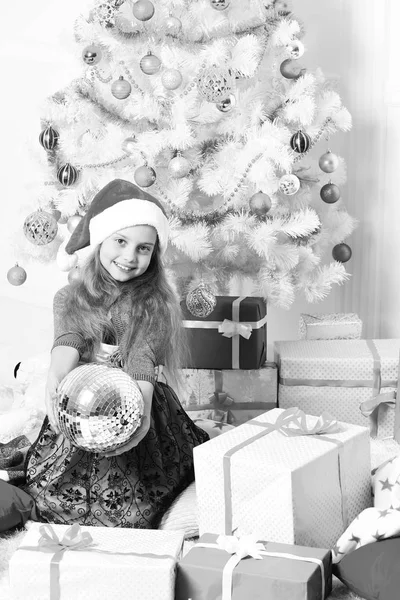 Christmas morning and New Year concept. Girl with happy face near white and blue Christmas tree on white background. Kid in Santa hat near present boxes. Little miss Santa holds disco ball