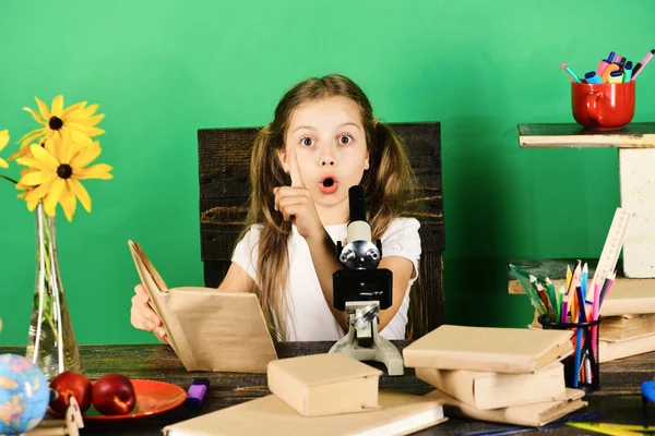 Školačka sedí u stolu s barevné kancelářské potřeby, knihy a mikroskop — Stock fotografie