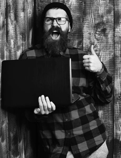 Bearded man, long beard. Brutal caucasian happy unshaven hipster holding laptop in red black checkered shirt with hat and glasses on brown vintage wooden studio backgroun
