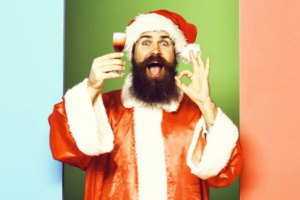 Knappe bebaarde kerstman met lange baard op een glimlachend gezicht met een glas alcoholische shot in rode kerst- of kerstjas en nieuwjaarshoed op een kleurrijke studioachtergrond — Stockfoto