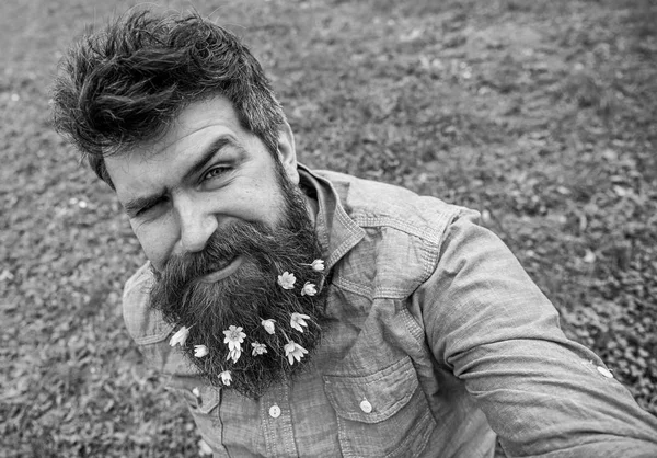 Hipster on winking face sits on grass, defocused. Man with beard enjoys spring, green meadow background. Guy with lesser celandine flowers in beard taking selfie photo. Spring season concept — Stock Photo, Image