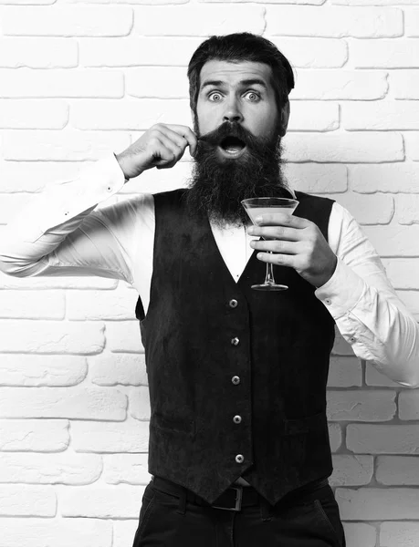 Bonito barbudo homem com longa barba e bigode tem cabelo elegante no rosto surpreso segurando vidro de bebida alcoólica em couro de camurça vintage colete o branco tijolo parede estúdio fundo — Fotografia de Stock