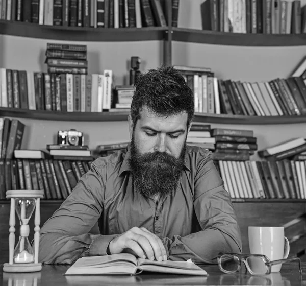 Hombre en el libro de lectura de la cara concentrada, estudio, estanterías en el fondo. Concepto de autoeducación. Profesor o estudiante con barba estudiando en la biblioteca. El hombre se sienta a la mesa con taza y reloj de arena —  Fotos de Stock