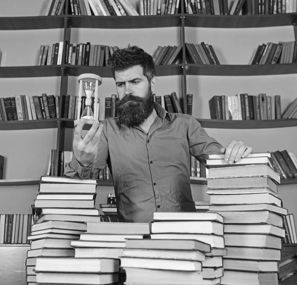 Conceptul bibliotecar. Profesor, om de știință cu barbă stă la masă cu cărți, deconcentrat. Omul pe o față grijulie stă între grămezi de cărți, în timp ce ține clepsidră, rafturi de cărți pe fundal — Fotografie, imagine de stoc