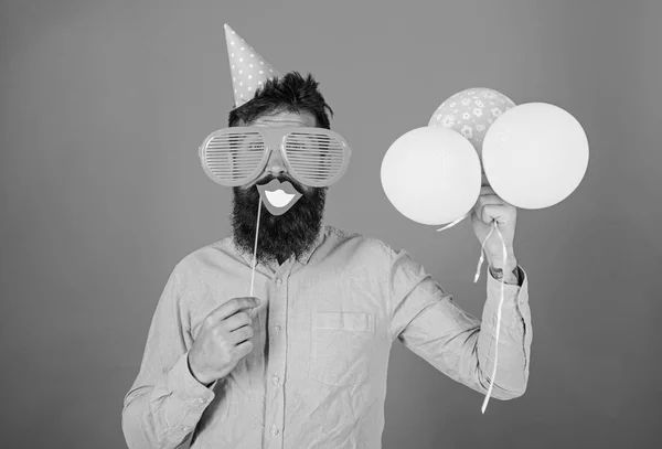 Hipster dans des lunettes de soleil géantes célébrant. Le gars en chapeau de fête avec des ballons à air célèbre. Homme avec barbe sur le visage joyeux tient la bouche souriante sur bâton, fond rouge. Concept amusant de photomaton — Photo