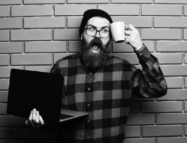 Sakallı, beyaz tenli hippi elinde bardakla dizüstü bilgisayar tutuyor.