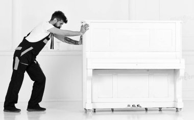 Man with beard and mustache, worker in overalls pushes piano, white background. Courier delivers furniture in case of move out, relocation. Courier service concept. Loader moves piano instrument clipart