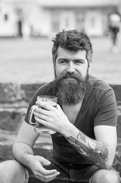 Hipster con barba larga se ve relajado. hipster barbudo sostiene taza de cerveza, bebe cerveza al aire libre. Concepto de cerveza artesanal. Hombre con barba y bigote en rostro sonriente, fondo pedregoso, desenfocado — Foto de Stock