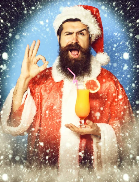Schöner bärtiger Weihnachtsmann mit langem Bart auf lustigem Gesicht, der ein Glas alkoholfreien Cocktails in Weihnachts- oder Weihnachtspullover und Neujahrsmütze auf blauem Studiohintergrund hält — Stockfoto