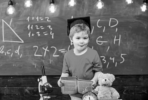 Πρώτη πρώην ενδιαφέρονται για σπουδές, εκπαίδευση. Παιδί αγόρι στο μεταπτυχιακό καπάκι κατέχει το βιβλίο στην τάξη, chalkboard σε φόντο. Παιδιού, μαθητή σχετικά με χαρούμενο πρόσωπο, κοντά στο μικροσκόπιο. Έννοια πια — Φωτογραφία Αρχείου