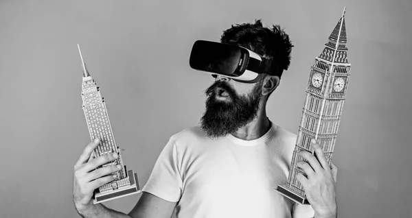 Man on excited face study architecture or design in virtual reality. 3D design concept. Guy in VR glasses holds Big Ben and Empire State Building. Man with beard in VR glasses, green background