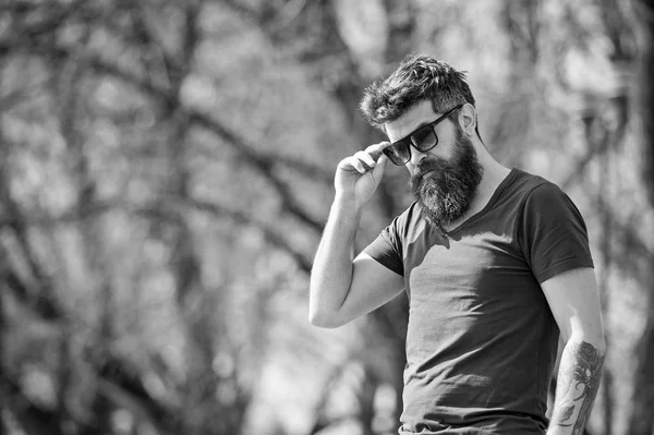 Hipster-Mann mit trendiger Sonnenbrille posiert auf einer Allee mit kahlen Bäumen. bärtiger Mann in dunkelblauem T-Shirt mit Tätowierung auf dem Arm Seitenansicht in der urbanen Szene an sonnigen Tagen — Stockfoto