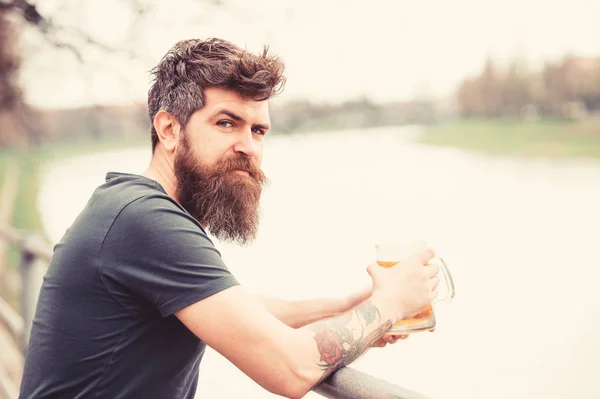 Man with long beard looks relaxed. Man with beard and mustache on calm face, river background, defocused. Bearded man holds beer mug, drinks beer outdoor. Craft beer concept