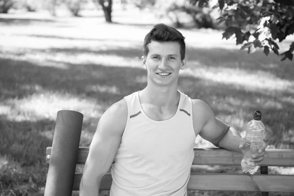 Hacia un estilo de vida más saludable. Hombre sonriendo cara con esterilla de yoga y botella de agua sentarse en el banco en el parque. Únete a la práctica de yoga al aire libre. Atleta con equipo de yoga relajante en el parque. El hombre eligió el yoga al aire libre — Foto de Stock